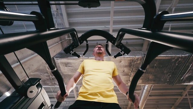 Man Performs Assisted Dips On Parallel Bars In The Gym With Legs Supported, Viewed From Below