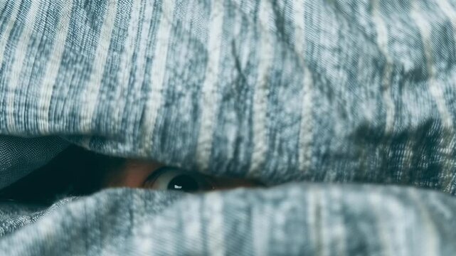 A man with wide open eyes is peeking out from under a blanket. Concept of curiosity and anticipation, as if the man is trying to uncover something hidden or mysterious. The blanket serves as a barrier