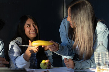 Two happy women sharing a banana and apple during a snack break, enjoying healthy food and cheerful...