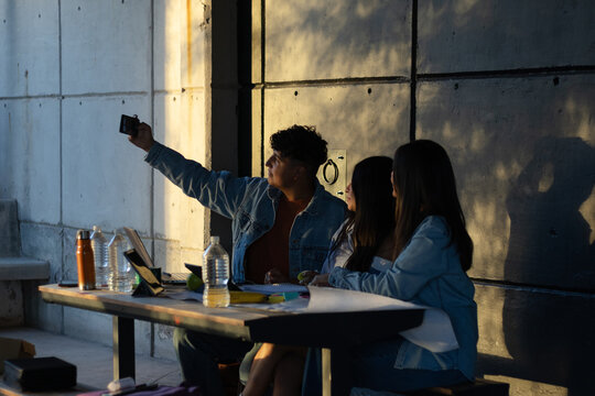 Diverse group of young adult students sitting outdoors at a table, taking a selfie together with a smartphone against a concrete wall during a warm sunset