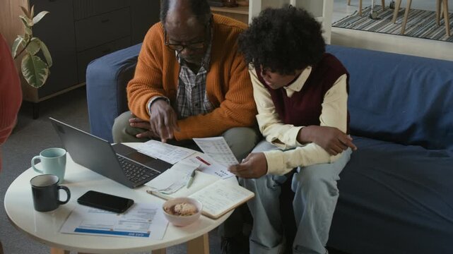 Focused elderly Black man reviewing receipts calculating utility payments with young grandsons at coffee table with open laptop in living room