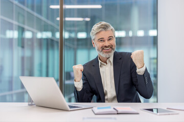 Happy senior businessman celebrating achievement at office desk, clenching fists in triumph and looking at the viewer with a beaming smile and excitement