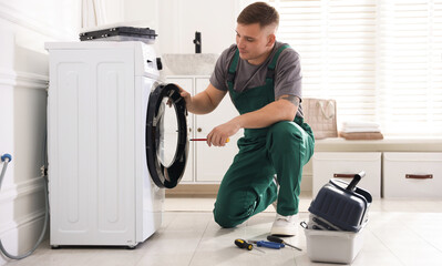 Repairman in uniform fixing broken washing machine with screwdriver at home