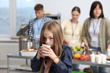 Volunteers giving food to homeless people in shelter, focus on girl with drink