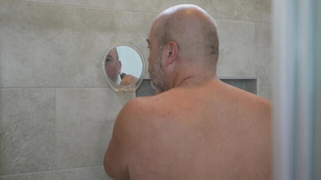 older man shaving his beard with a small mirror in the shower