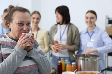 Volunteers giving food to homeless people in shelter, focus on senior woman with drink