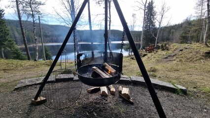 Scenic outdoor cooking setup by a peaceful lake in the forest with burning firewood during daytime