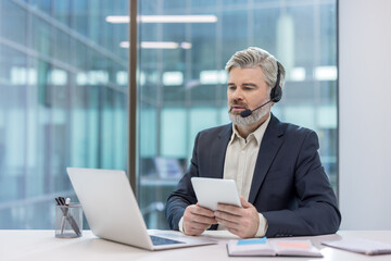 Senior businessman wearing a headset and holding a tablet, providing online customer support or engaging in a video call at his modern corporate office desk