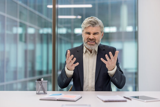 Senior businessman displaying intense emotions of frustration and anger, gesturing with open hands while sitting at his desk in a modern office environment during a stressful business interaction - Powered by Adobe