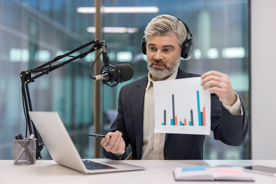 Man in headphones and suit jacket creating a podcast, presenting a report with a bar chart and statistics, using a studio microphone and laptop during an online webinar