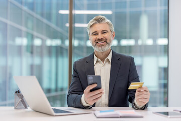 Mature businessman smiling at camera, holding a credit card and smartphone while sitting at an office desk with a laptop, engaging in e-commerce and mobile banking