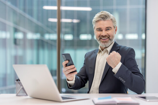 Mature businessman receiving good news on his smartphone, smiling and clenching a fist in celebration of a victory or achievement in a modern office environment