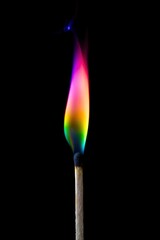 A single wooden matchstick ignites with a breathtakingly colorful flame, radiating a vibrant spectrum of hues against a stark, deep black background. The mesmerizing fire displays a beautiful gradient