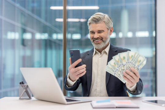 Mature businessman in modern office holding a stack of hundred-dollar bills and a smartphone, smiling confidently after a successful digital banking transaction and profit realization