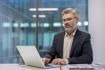 Professional mature man with a beard and glasses using a laptop while sitting at a desk in a contemporary office setting, performing online business tasks