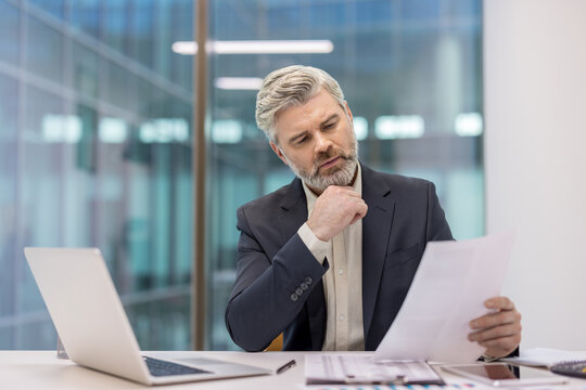 Mature businessman with gray hair and beard concentrating on a document, sitting at a modern office desk with a laptop and papers, representing focus and analysis in a corporate environment
