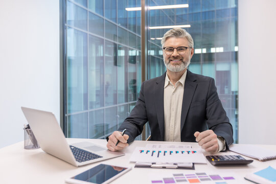 Experienced male entrepreneur with gray beard wearing glasses, sitting at an office desk with a laptop, tablet, and financial charts, confidently planning business strategies