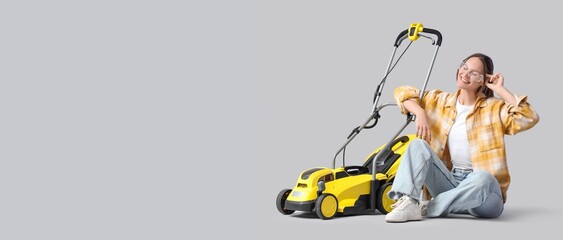 Young woman sitting near modern lawn mower on grey background with space for text