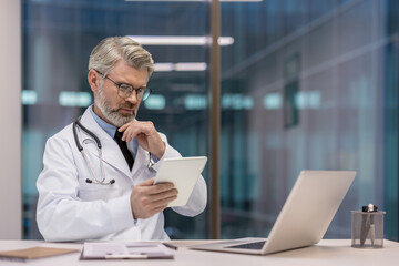 Mature male doctor wearing a white lab coat and stethoscope, sitting at a desk and reviewing healthcare information on a tablet in a modern office environment