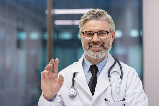 Experienced male doctor wearing a white coat, stethoscope, and glasses, smiling confidently and providing reassurance with an okay gesture from a clinic or hospital setting