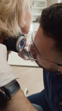 Vertical shot of male doctor in white gloves examining skin condition on neck of blonde female patient using dermatoscope during consultation in clinic