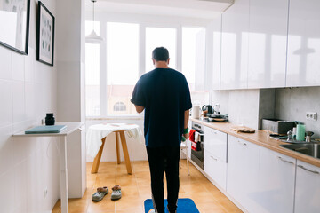 person monitoring body weight in bright kitchen, man using scale for fitness tracking near window light