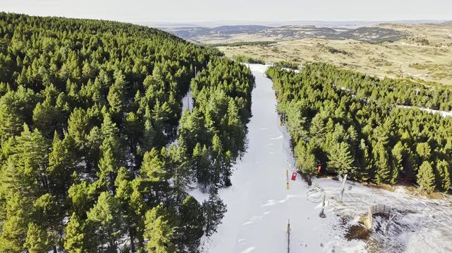 Drone flight parallel to a snowy mountain ski slope surrounded by dense pine forest, winter landscape, alpine terrain, ski resort infrastructure, peaceful nature, scenic aerial motion, winter tourism