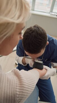 Vertical high angle shot of young male doctor in scrubs examining arm of blonde female patient using dermatoscope during medical appointment in clinic