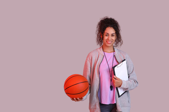 Female African-American basketball trainer with ball and clipboard on pink background