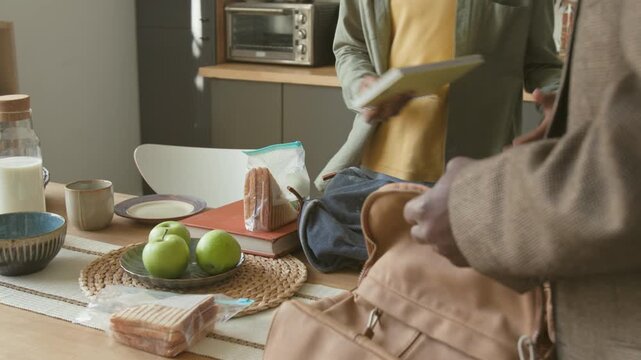 Senior African American man helping two young grandsons packing backpacks with healthy lunch and snacks at kitchen table while siblings leaving for school in morning