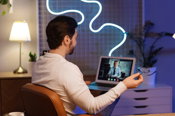 Handsome man with laptop video chatting in office at night, back view