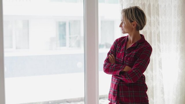 Young woman in red plaid pajamas opens the window, letting in fresh air. Her posture reflects relief and renewed energy. Concept of recovery, wellness, and fresh start. Back view, natural light, mid