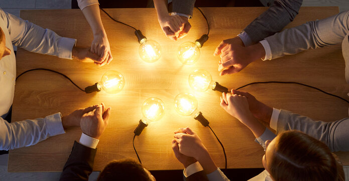 Cropped top view on group of business professionals sitting around wooden desk, holding hands in circle with glowing light bulbs between them. Concept of teamwork, innovation and unity. Banner.