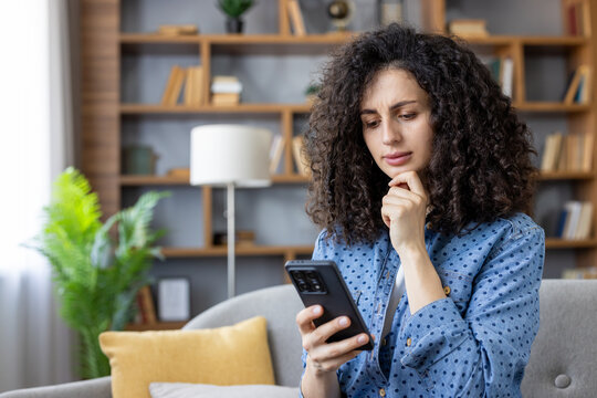 Young woman with curly hair sitting on a couch at home, focusing on a smartphone screen with a concerned expression, contemplating a difficult message or shocking news