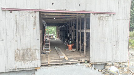 Construction workers are renovating a barn, with wooden beams, scaffolding, and tools visible. The setting is a rural area, showing the rebuilding process of an old structure.