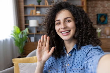 Young curly-haired woman waving hand, smiling, communicating through a video call or videoconference from home, engaging in virtual meeting or online conversation, looking at camera comfortably