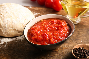 Board with raw dough and bowl of tasty tomato sauce, closeup