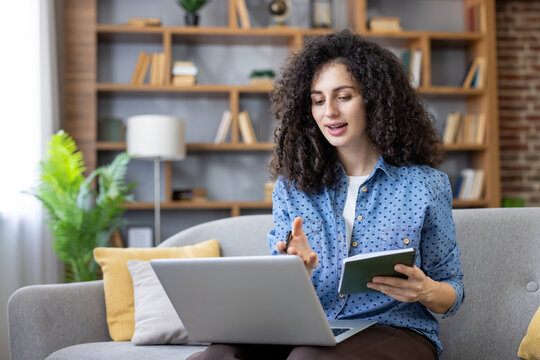 Young woman with curly hair engaging in an online video call or learning session on her laptop, sitting comfortably on a sofa in her home office setup