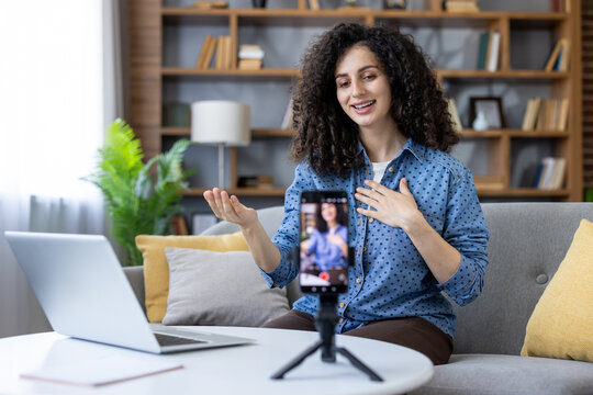 Woman creating online content, talking to camera during a successful livestream or video call from home, using a smartphone on a tripod with a laptop present