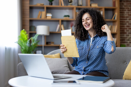 Happy young woman celebrating exciting news, opening an acceptance letter with a fist pump, sitting on a sofa with a laptop at home or in a student dormitory