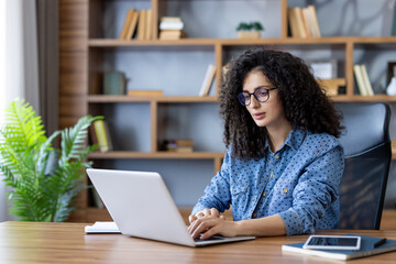 Young woman with curly hair and glasses concentrating on her work, typing on a laptop at a wooden desk in a modern home office with a bookshelf in the background