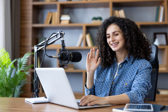 Young woman smiling and waving at laptop screen during a video call, livestream, or podcast recording from her home office with a professional microphone - Powered by Adobe
