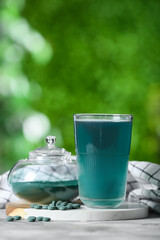 Glass of spirulina drink with pills, jar and powder on grey grunge table outdoors