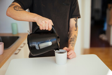 Tattoed individual gently pours hot water into mug during morning routine