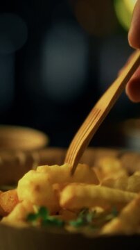 A man dips crispy French fries into a creamy sauce using wooden cutlery. This eco-friendly choice highlights a sustainable dining option in a cozy setting, perfect for casual meals