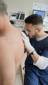 Vertical shot of focused young Middle Eastern male dermatologist in scrubs and white gloves using dermatoscope while examining skin condition of unrecognizable male patient in clinic