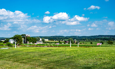 Obraz premium A wide view of a peaceful rural area showing green fields with white fences and American flags. Farms dot the landscape under a bright blue sky filled with fluffy clouds.