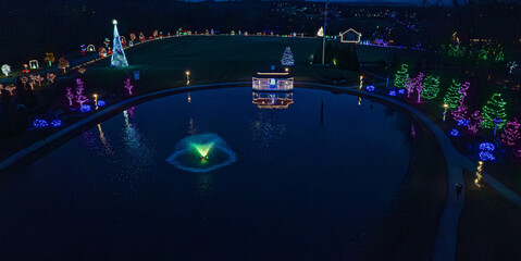 Colorful holiday lights brighten a park pond at night, showcasing decorated trees and structures around the water. The scene captures festive cheer and joy during the holiday season.