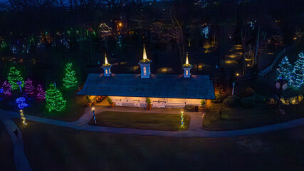 Brightly lit trees surround a quaint pavilion filled with holiday decorations. People stroll through the park, enjoying the beautiful light displays as night falls.
