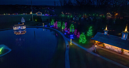 People enjoy a holiday light display at a park at night. Vibrant colors decorate trees and buildings around a peaceful pond, creating a festive atmosphere for visitors to explore.
